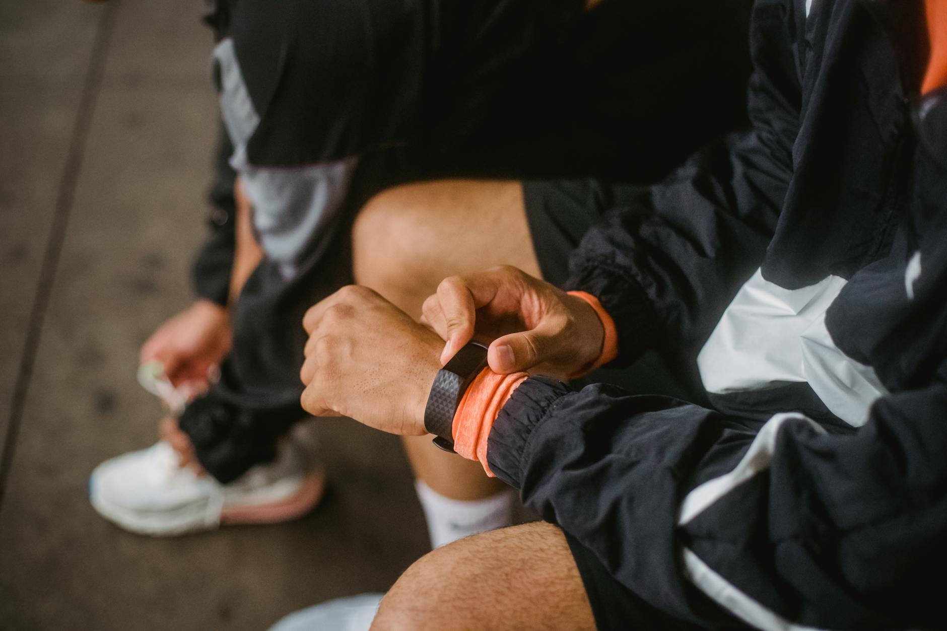 Homem sentado, olhando atentamente o relógio de corrida, refletindo sobre seu desempenho enquanto se prepara para a próxima etapa do treino ou competição. Ao seu lado, outra pessoa está ajudando a amarrar seu tênis,